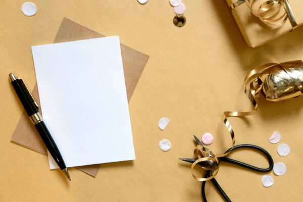 Flat lay of a blank card, pen, scissors, and gift-wrapping items on a brown surface.